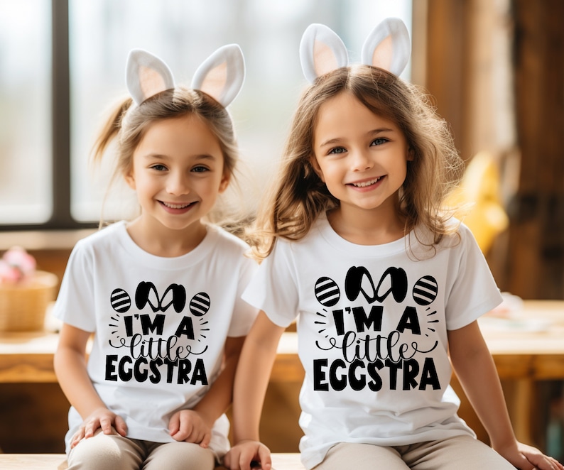 May include: Two children wearing white t-shirts with black bunny ears and the text "I'm a little eggstra" printed on them. The children are smiling and looking at the camera.