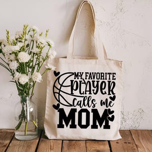 May include: A white canvas tote bag with black text that reads "My Favorite Player Calls Me Mom". A basketball graphic is next to the text. There are small hearts around the text and graphic.