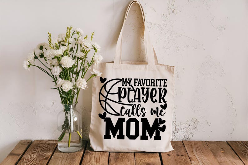 May include: A white canvas tote bag with black text that reads "My Favorite Player Calls Me Mom". A basketball graphic is next to the text. There are small hearts around the text and graphic.