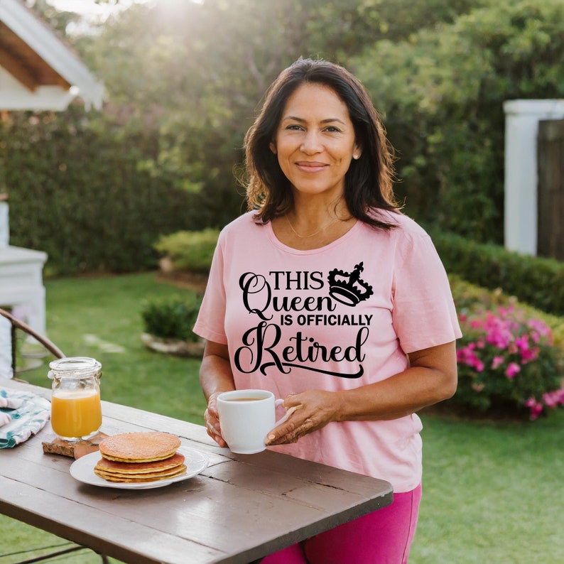May include: A woman wearing a pink t-shirt with the text "This Queen is Officially Retired" and a crown graphic. She is holding a white mug and smiling.