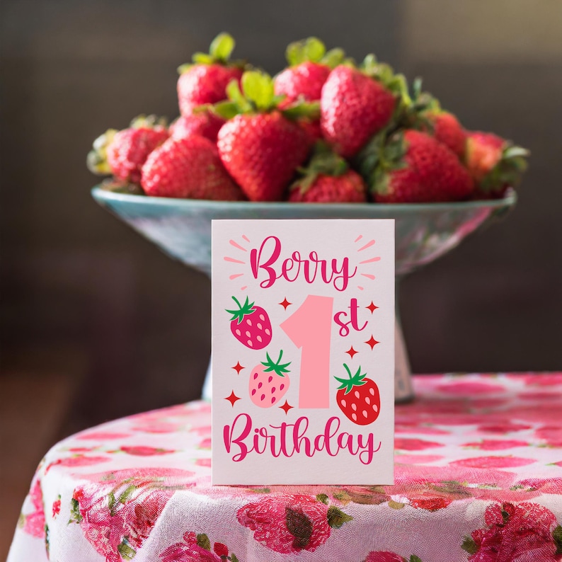 May include: A pink and white card with the text "Berry 1st Birthday" and a pink number one surrounded by strawberries. The card is sitting on a pink and white floral tablecloth.