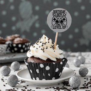May include: A chocolate cupcake with white frosting and black sprinkles topped with a silver round cake topper that says "Birthday Boy Time to Level Up" with a black and white video game controller graphic.