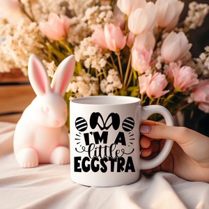 May include: White ceramic mug with black text that reads "I'm a little eggstra" and a graphic of two Easter eggs. The mug is sitting on a white surface with a pink bunny rabbit to the left.
