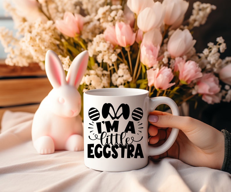 May include: White ceramic mug with black text that reads "I'm a little eggstra" and a graphic of two Easter eggs. The mug is sitting on a white surface with a pink bunny rabbit to the left.