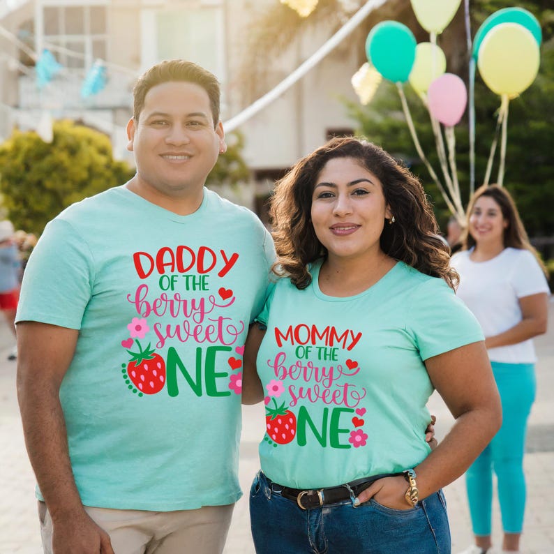 Puede incluir: Una pareja lleva camisetas azules claras a juego con el texto "Daddy of the berry sweet one" y "Mommy of the berry sweet one" en letras rojas y verdes. Las camisetas presentan un gr&aacute;fico de fresa con flores rosas.