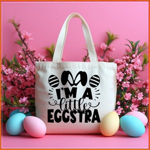 May include: A white canvas tote bag with black text that reads "I'm a little eggstra". The bag is surrounded by pastel-colored Easter eggs and pink flowers.