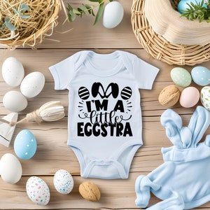 May include: A light blue baby onesie with the text "I'm a little eggstra" in black. The onesie is surrounded by Easter eggs in various colors and patterns.