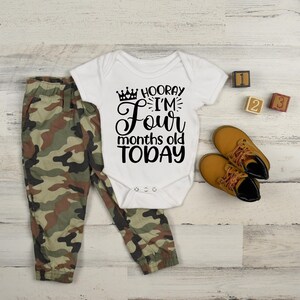 May include: A white bodysuit with black text that reads 'Hooray I'm Four months old today' with a crown graphic. The bodysuit is on a white wooden surface with a pair of brown leather shoes, a pair of camouflage pants, and three wooden blocks with the numbers 1, 2, and 3.