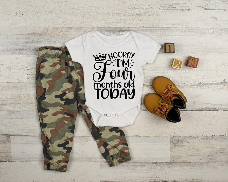 May include: A white bodysuit with black text that reads 'Hooray I'm Four months old today' with a crown graphic. The bodysuit is on a white wooden surface with a pair of brown leather shoes, a pair of camouflage pants, and three wooden blocks with the numbers 1, 2, and 3.