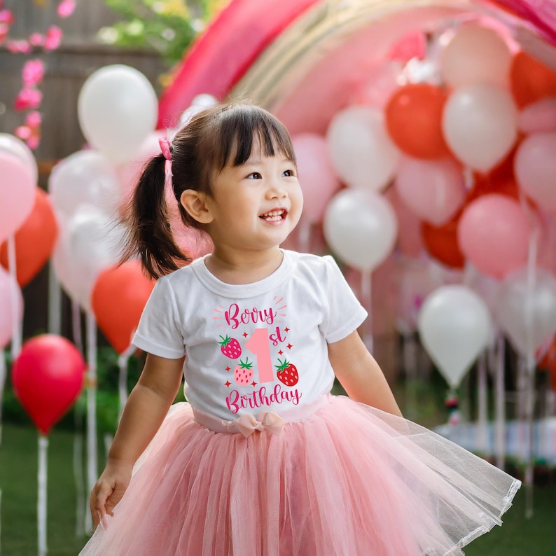May include: A young girl wearing a white t-shirt with a pink strawberry design and the text "Berry 1st Birthday". She is wearing a pink tulle skirt and is surrounded by pink and white balloons.