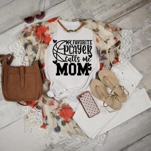 May include: White t-shirt with a black basketball graphic and the text "My Favorite Player Calls Me Mom". The shirt is paired with a white lace cardigan, white jeans, and brown leather purse.
