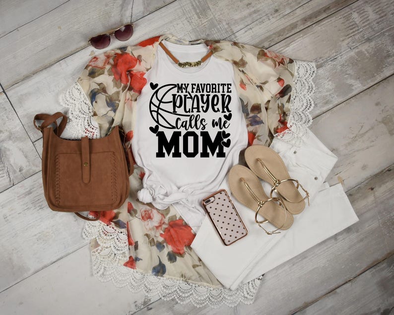 May include: White t-shirt with a black basketball graphic and the text "My Favorite Player Calls Me Mom". The shirt is paired with a white lace cardigan, white jeans, and brown leather purse.