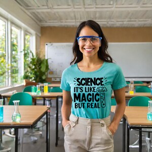 May include: A woman wearing a light blue t-shirt with the text "Science it's like magic but real" in black lettering. She is wearing safety glasses and standing in a classroom.