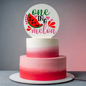 May include: A two-tiered pink and white cake with a white circle topper that says "one in a melon". The topper features a red watermelon slice with green seeds.