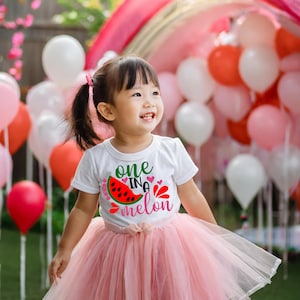 May include: A young child wearing a white t-shirt with a watermelon graphic and the text "one in a melon" in pink and green. The child is wearing a pink tulle skirt and is surrounded by pink and white balloons.