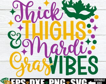 Puede incluir: Texto morado, verde y amarillo sobre un fondo de madera blanca que dice "Thick Thighs & Mardi Gras Vibes". Una máscara verde de Mardi Gras está sobre el texto.
