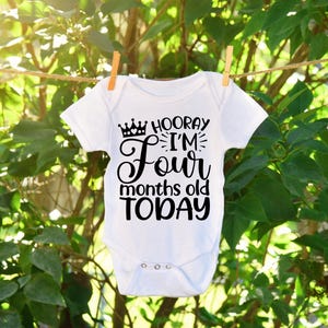 May include: A white baby onesie with black text that reads 'Hooray I'm Four months old TODAY' with a crown above the word 'Hooray'.