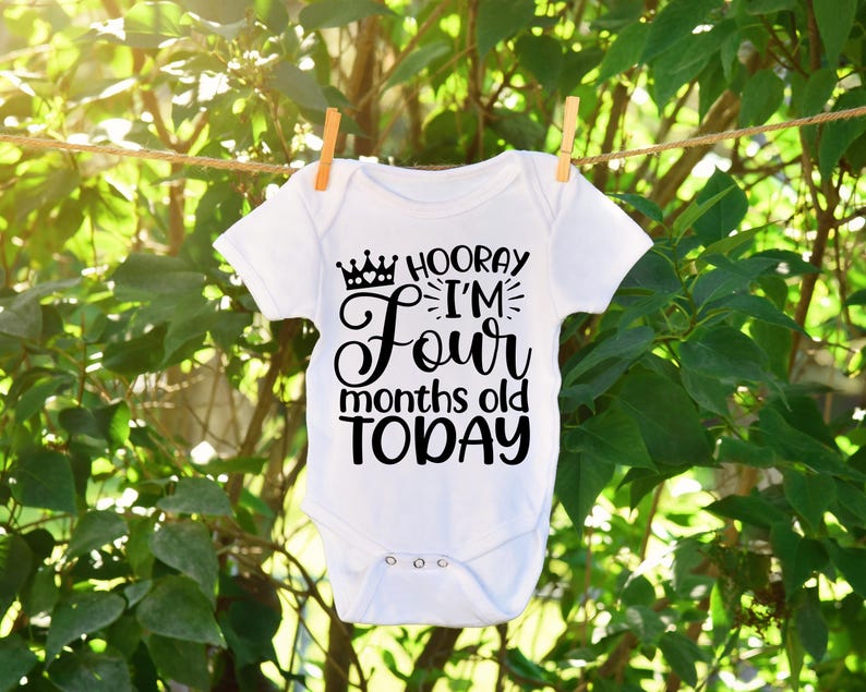 May include: A white baby onesie with black text that reads 'Hooray I'm Four months old TODAY' with a crown above the word 'Hooray'.