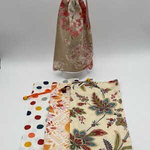 May include: A collection of drawstring bags in various floral and patterned designs. One tall bag features a beige background with pink floral patterns. Three flat bags display polka dots, autumn leaves, and a cream floral print. Each bag has a drawstring closure.