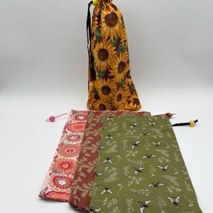 May include: Three fabric drawstring bags with different patterns. The first bag is pink and white with a floral pattern. The second bag is brown with a leaf pattern. The third bag is green with a bee and leaf pattern.