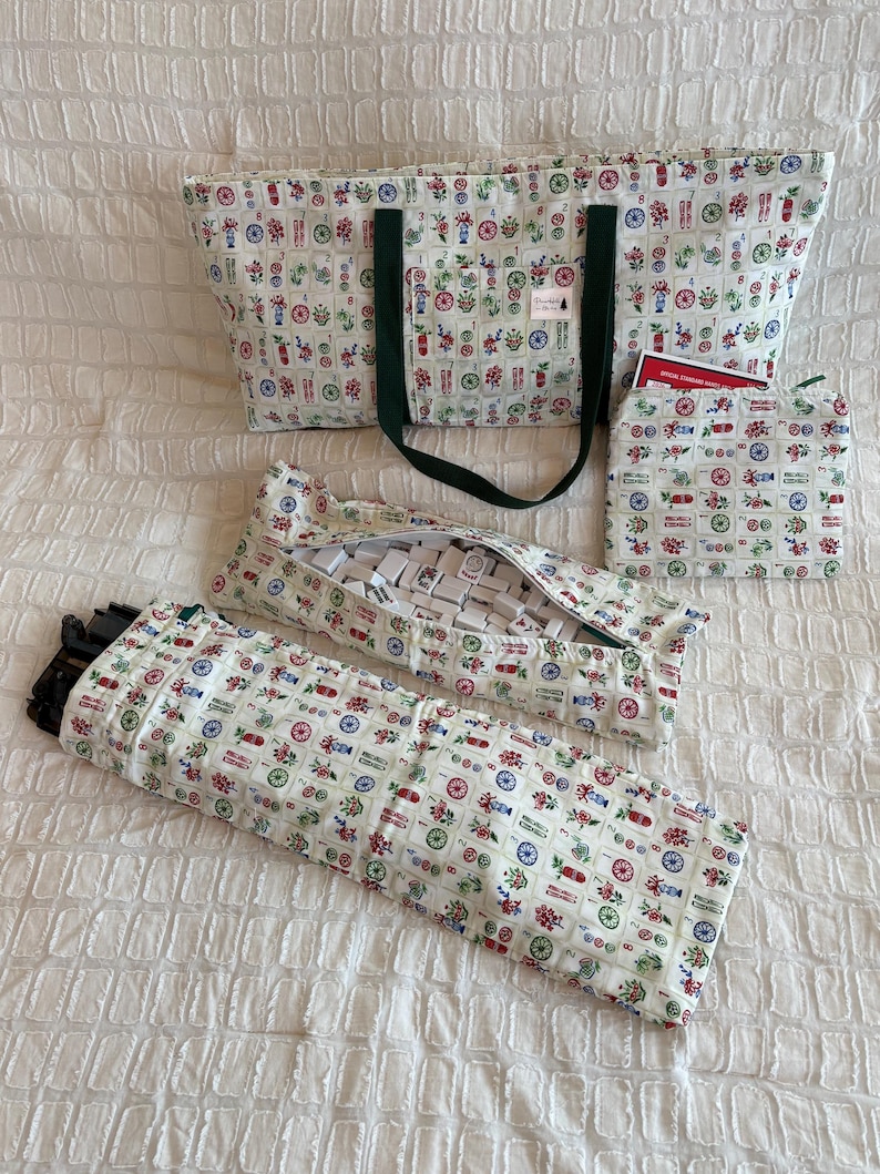 May include: A set of Mahjong game accessories, including a tote bag, tile cases, and a small pouch. The items are made of a cream-colored fabric with a repeating pattern of colorful floral and geometric designs. The tote bag has dark green handles.