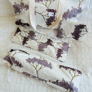 May include: A set of four fabric items featuring a cream background with a dark purple tree pattern. The collection includes a large tote bag with white straps and a small label, a rectangular zippered pouch, a long flat case, and a cylindrical bag.