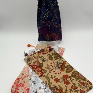 May include: Three fabric drawstring bags with different patterns. One bag has a navy blue background with gold constellations, another has a beige background with red and orange floral designs, and the third has a beige background with pink and white floral designs.