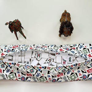 May include: A zippered fabric case filled with white Mahjong tiles, with a colorful pattern of red, blue, and green symbols. Two small, decorative animal figurines are positioned above the case.