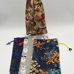 May include: Four fabric drawstring bags with different patterns. One bag has a floral pattern in shades of brown and red. Another bag has a navy blue background with orange and green floral accents. The third bag has a gray background with a white dog wearing sunglasses pattern. The last bag has a dark purple background with a constellation pattern.