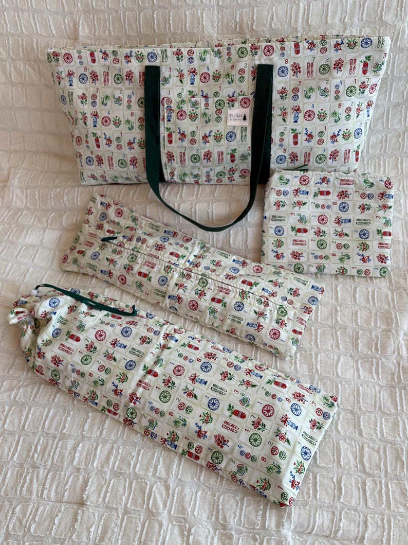 May include: A set of matching fabric items, including a tote bag, a small square pouch, and two long, cylindrical pouches. The items are white with a repeating pattern of colorful floral and geometric designs. The tote bag has dark green handles.