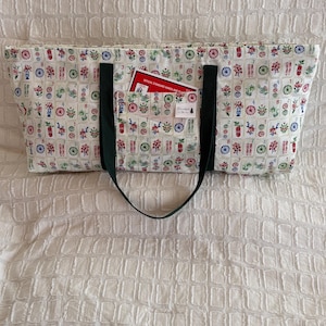 May include: A rectangular tote bag with a white background and a colorful pattern of flowers and game tiles. The bag has a dark green strap and a pocket with a red card that reads "OFFICIAL STANDARD HANDS AND RULES".