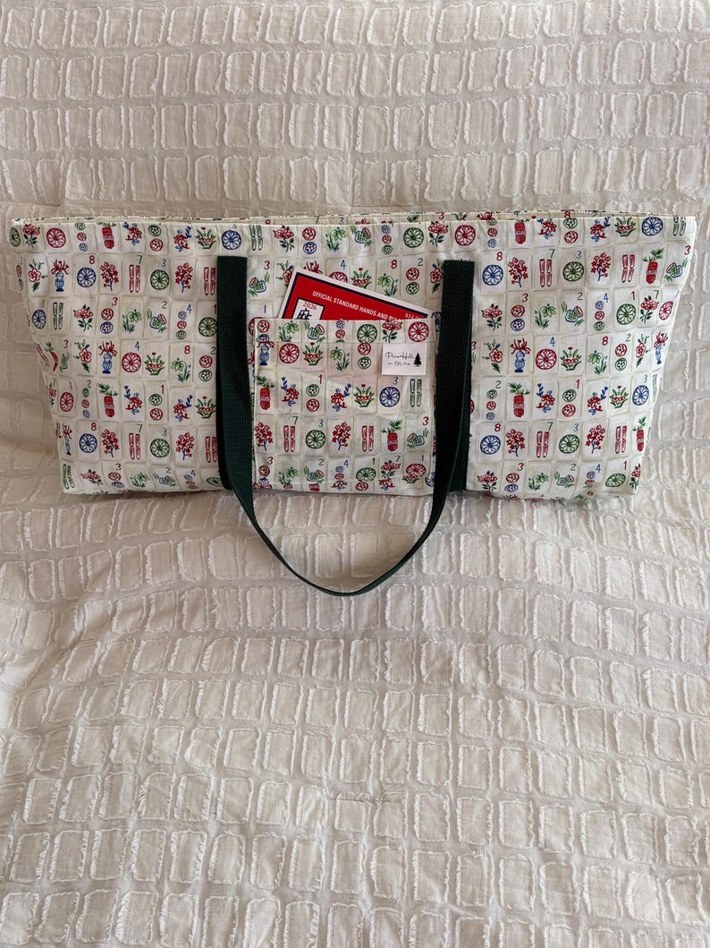 May include: A rectangular tote bag with a white background and a colorful pattern of flowers and game tiles. The bag has a dark green strap and a pocket with a red card that reads "OFFICIAL STANDARD HANDS AND RULES".