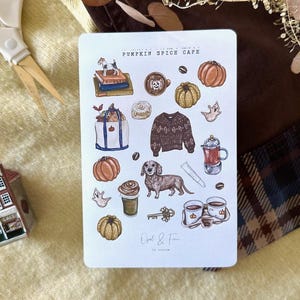 May include: A white sticker sheet with illustrations of pumpkins, coffee, a dachshund, and a sweater. The text "Pumpkin Spice Cafe" is at the top. A miniature house and scissors are in the background.