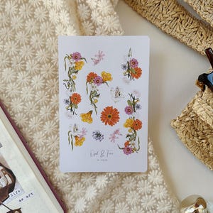 May include: A white card featuring floral illustrations in orange, yellow, pink, and purple. The card is placed on a cream-colored, textured surface, next to an open journal with collaged images and a woven bag with sunglasses.