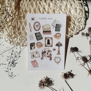 Puede incluir: Una hoja de pegatinas con ilustraciones de libros, tazas de café, un perro, una lámpara y un suéter. Las pegatinas se titulan "A Library Date" y son de "Owl & Fox".