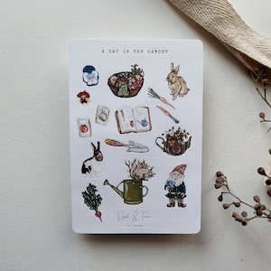 May include: A sheet of stickers with a garden theme. The stickers feature a bunny rabbit, a gnome, a watering can, a basket of vegetables, a book, a trowel, a flower pot, and a few other garden-related items. The stickers are printed on white paper and have a whimsical, hand-drawn style.