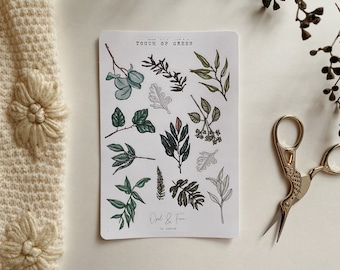 May include: A sticker sheet with botanical illustrations of various green leaves and branches. The text "Touch of Green" is at the top. Gold scissors and a cream-colored floral textile are also in the image.