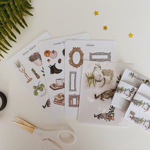 May include: A collection of decorative stickers and stationery featuring botanical and autumnal themes. The stickers include illustrations of plants, frames, cats, and seasonal elements. Also included are washi tape rolls and a pair of scissors.