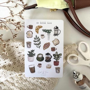 May include: A sticker sheet with cafe-themed illustrations, including coffee cups, croissants, plants, and a French press. The text "My Local Cafe" is at the top. The sheet is surrounded by a brown purse, scissors, and a perfume bottle.
