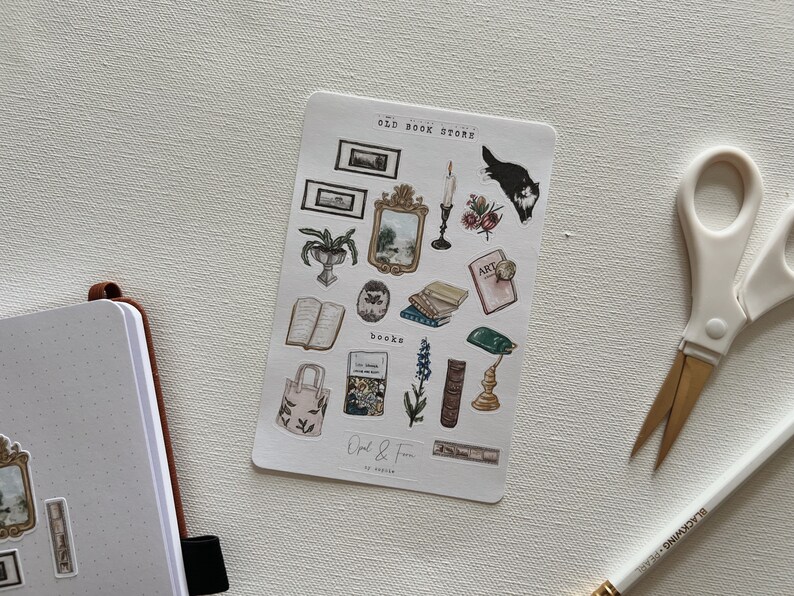 May include: A sticker sheet with illustrations of vintage books, mirrors, candles, and a black and white cat. The sheet is labeled "OLD BOOK STORE" and includes the text "Opal & Fern". A notebook, scissors, and pencil are in the background.