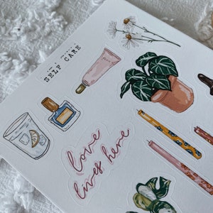 Peut inclure: Une feuille d'autocollants sur le th&egrave;me du bien-&ecirc;tre. Les autocollants pr&eacute;sentent des illustrations d'un verre d'eau avec un quartier de citron, un flacon de parfum, un tube de lotion, une plante en pot, des bougies et un flacon d'huile essentielle. Le texte "love, live, here" est &eacute;crit en cursive rose.