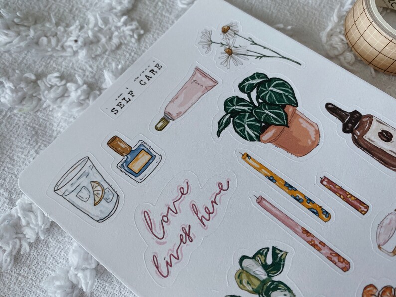 Peut inclure: Une feuille d'autocollants sur le th&egrave;me du bien-&ecirc;tre. Les autocollants pr&eacute;sentent des illustrations d'un verre d'eau avec un quartier de citron, un flacon de parfum, un tube de lotion, une plante en pot, des bougies et un flacon d'huile essentielle. Le texte "love, live, here" est &eacute;crit en cursive rose.