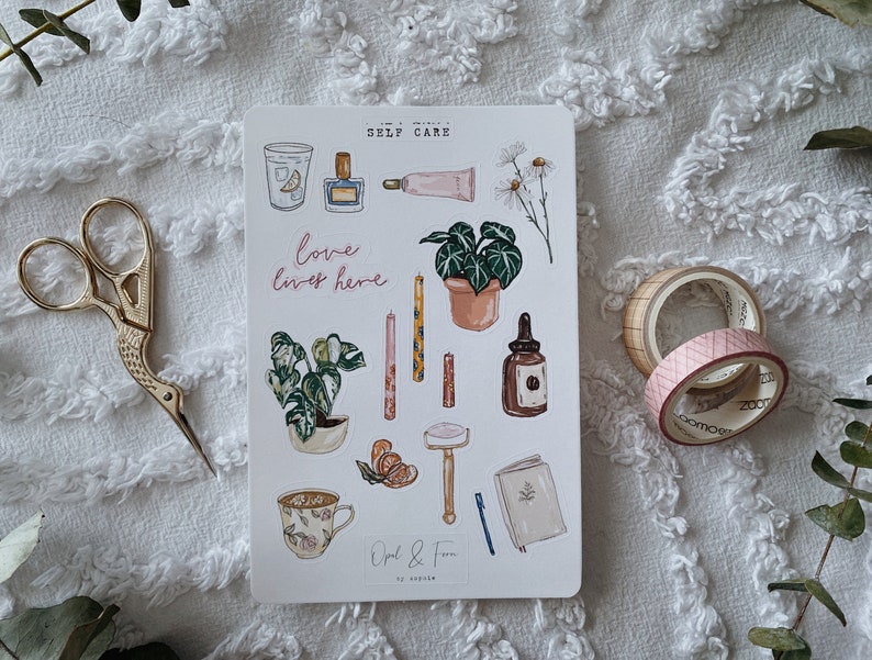 Peut inclure: Une feuille d'autocollants illustrant des articles de soins personnels, notamment des plantes, des bougies, un verre d'eau avec du citron, un flacon de parfum, un tube de cr&egrave;me pour les mains, un rasoir, un carnet, une tasse de th&eacute; et une bouteille d'huile essentielle. Les autocollants sont sur un fond blanc et comportent le texte "Self Care" et "Love lives here" en rose.