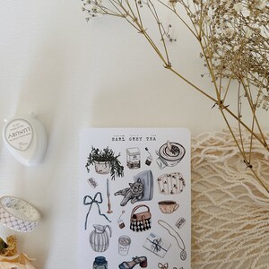 May include: A sheet of stickers with illustrations of teacups, teapots, a cat, a potted plant, and other tea-related items. The stickers are on a white background and have a vintage aesthetic. The text "Earl Grey Tea" is printed at the top of the sheet.