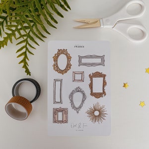 May include: A sticker sheet featuring various frame designs in gold and brown tones, with the text "FRAMES" at the top. The image includes washi tape, scissors, fern leaves, and gold star-shaped confetti.