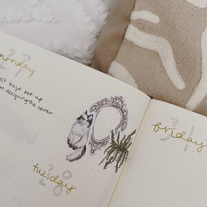 May include: Open journal with gold lettering for days of the week. A decorative sticker of a cat, ornate frame, and plant are on the left page. The journal is open on a bed with a white and tan pillow.