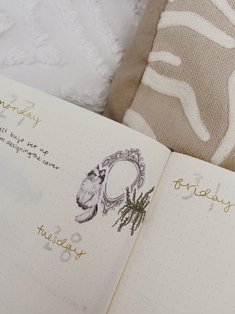 May include: Open journal with gold lettering for days of the week. A decorative sticker of a cat, ornate frame, and plant are on the left page. The journal is open on a bed with a white and tan pillow.