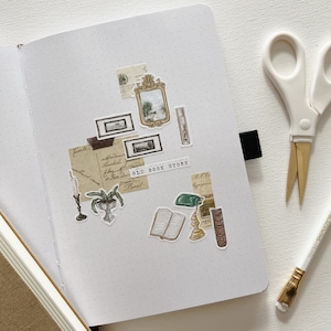 May include: An open dot grid journal with vintage-themed stickers, including framed art, an open book, and a candle. The stickers are arranged on the right page, with the text "OLD BOOK STORE". A pair of scissors and a pencil are on the right.