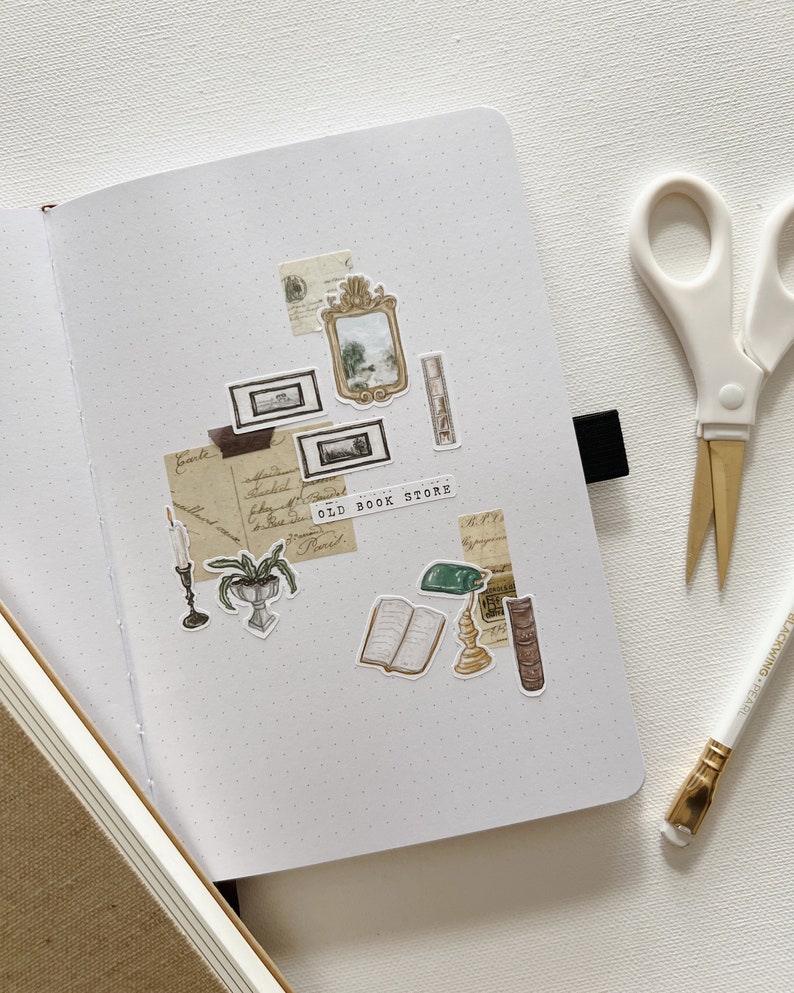 May include: An open dot grid journal with vintage-themed stickers, including framed art, an open book, and a candle. The stickers are arranged on the right page, with the text "OLD BOOK STORE". A pair of scissors and a pencil are on the right.