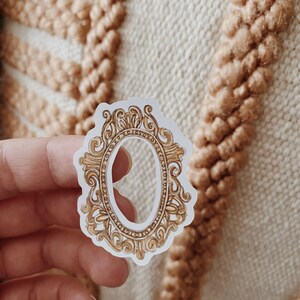 May include: A sticker featuring a gold-colored, ornate oval frame design. The frame has intricate scrollwork and detailing, with a white background. The sticker is held in a hand against a textured, beige background.